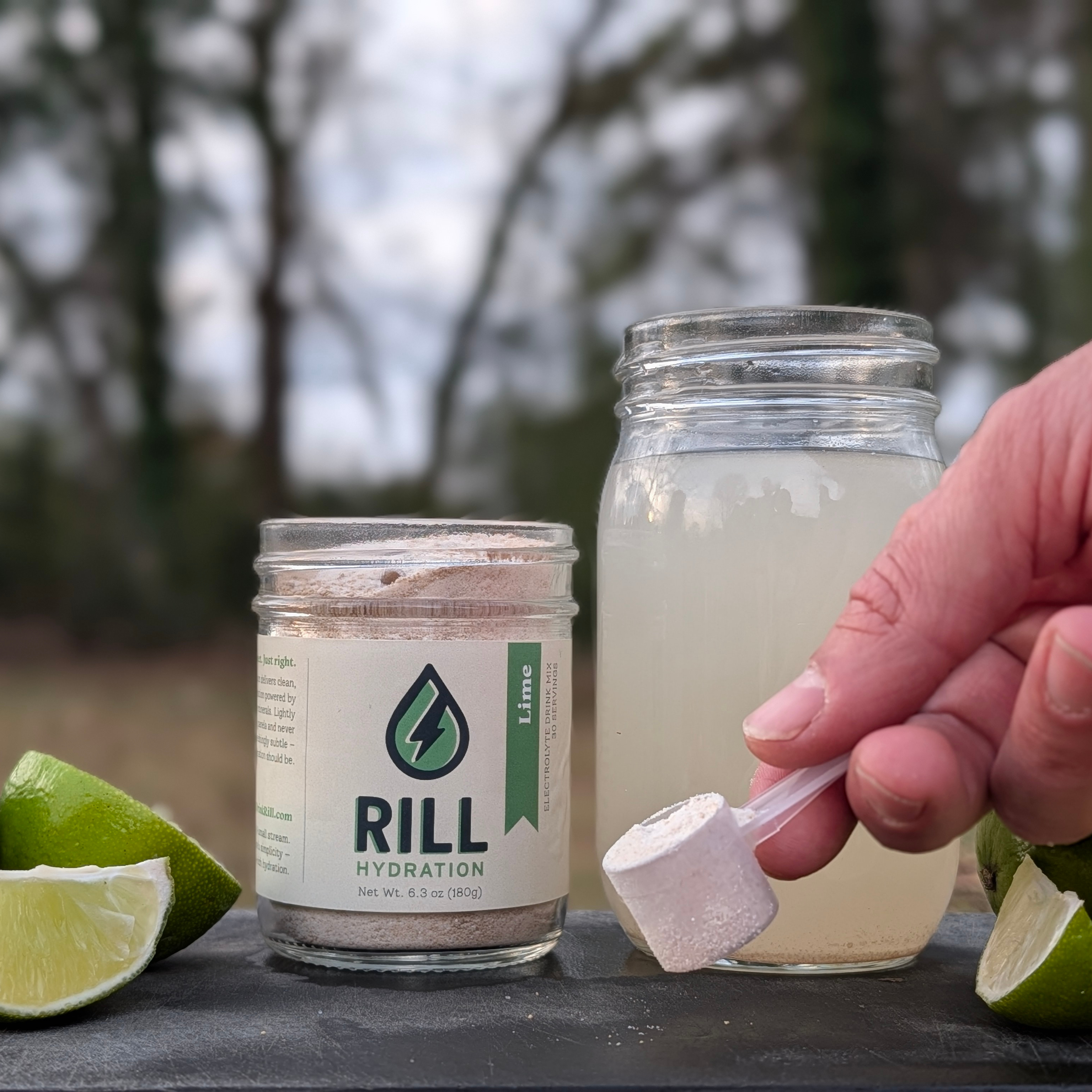 RILL Hydration product with a jar of lime-flavored electrolyte drink mix 
and a hand holding a scoop outdoors.
