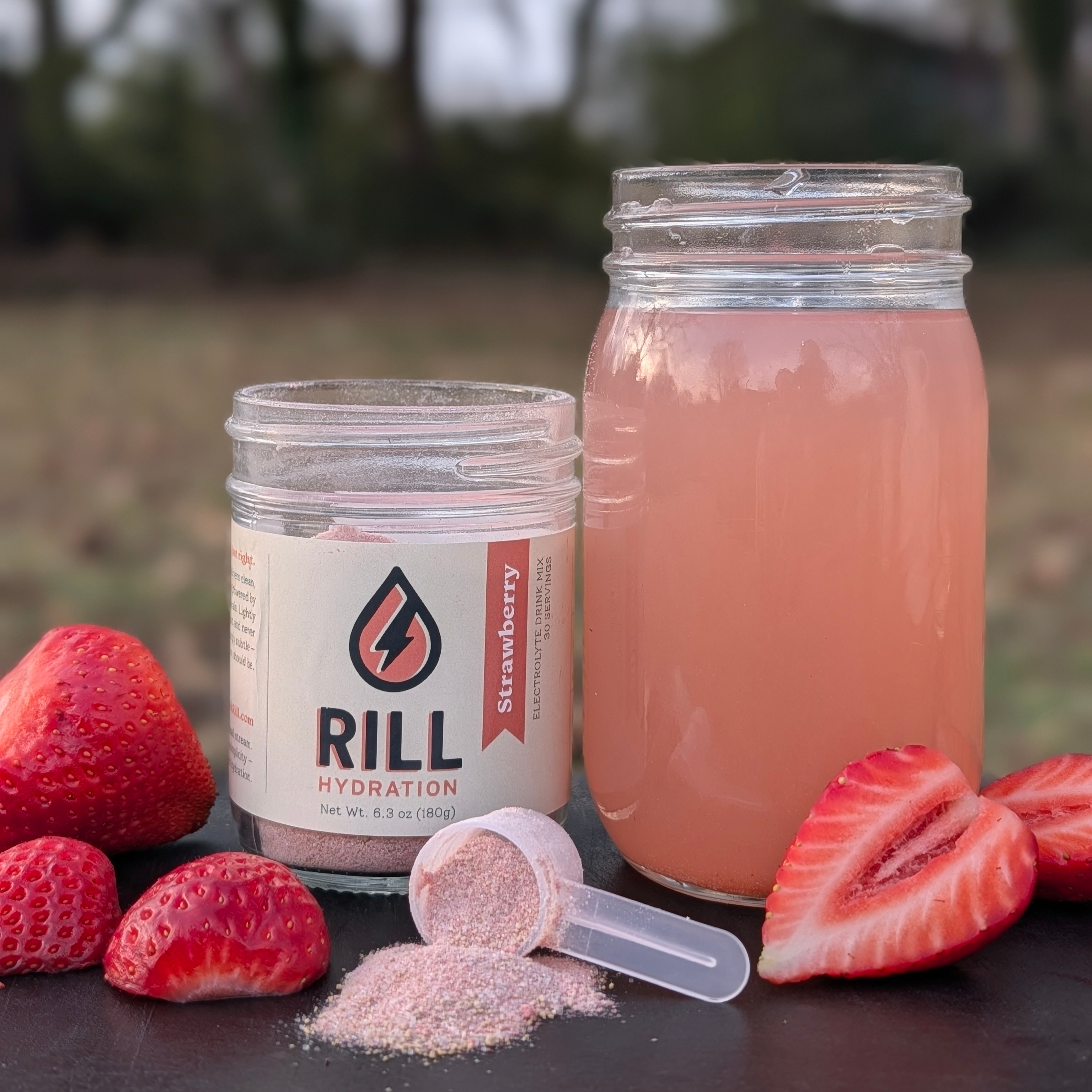RILL hydration strawberry electrolyte drink mix powder with a jar full of drink and fresh strawberries outdoors.