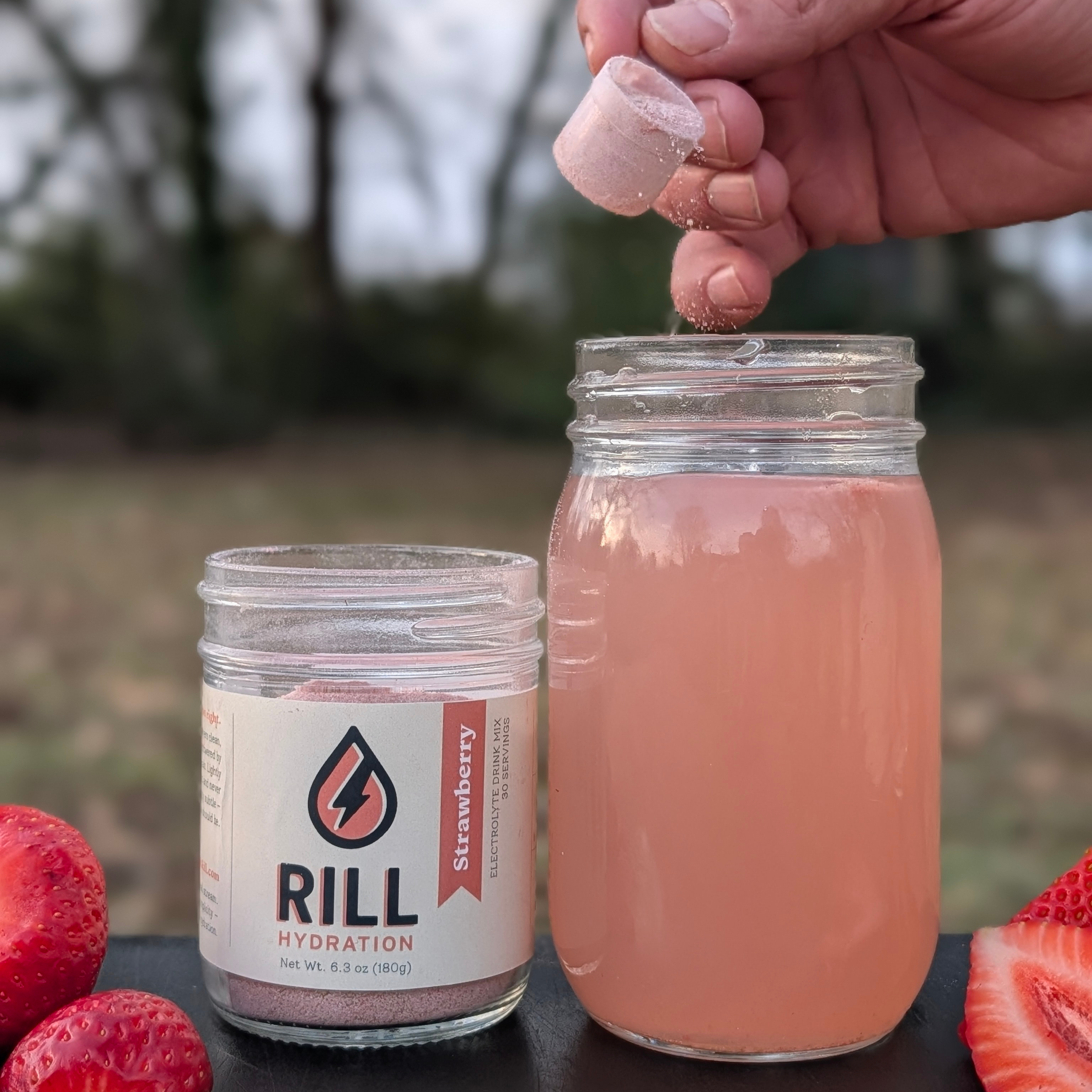 Person adding a RILL Hydration strawberry electrolyte drink mix powder to a glass of pink liquid with strawberries around.
