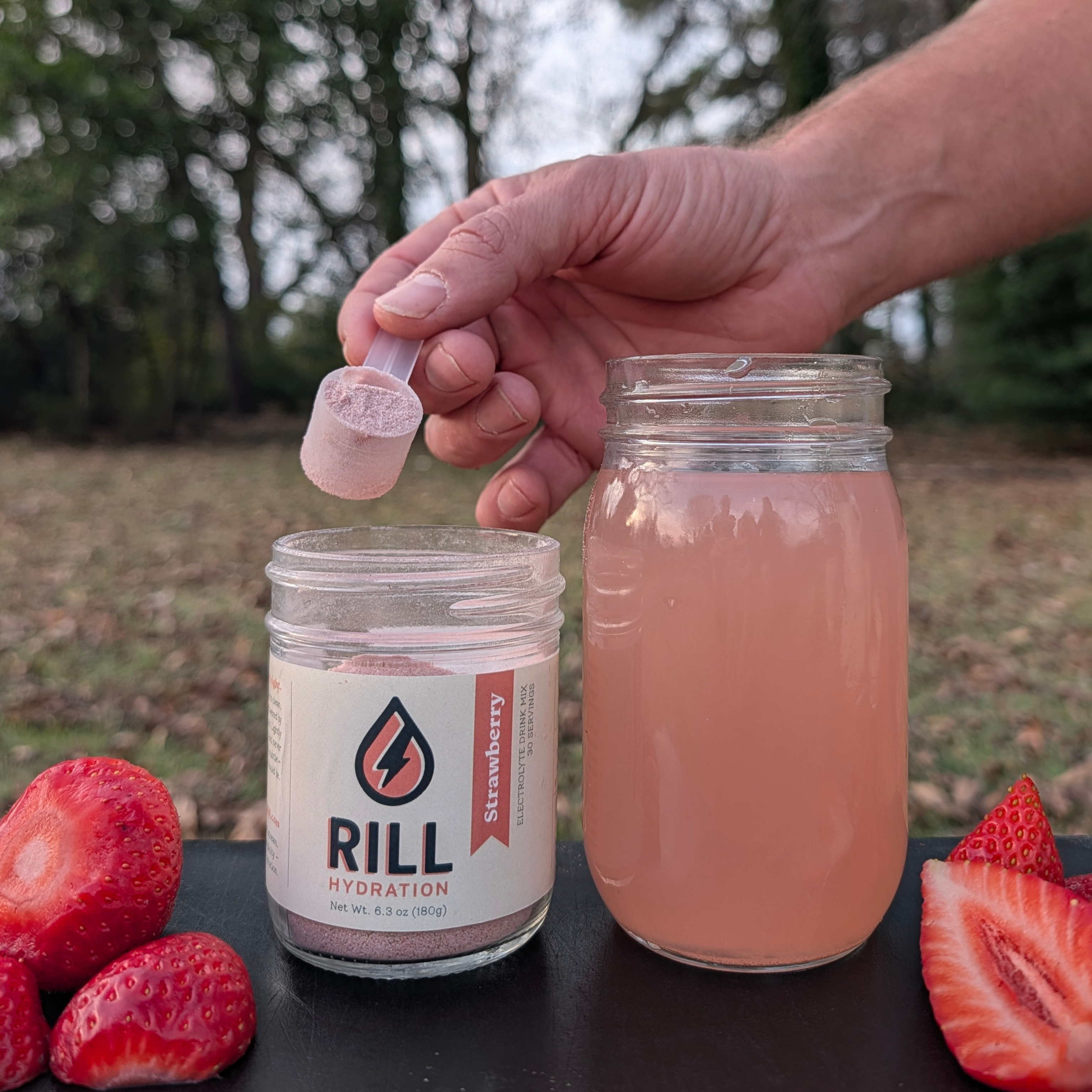 Person using a RILL Hydration Strawberry electrolyte drink mix, surrounded by strawberries outdoors.